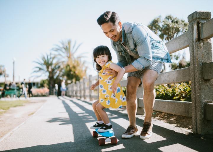 A child is riding a skateboard while being pushed along by her father.